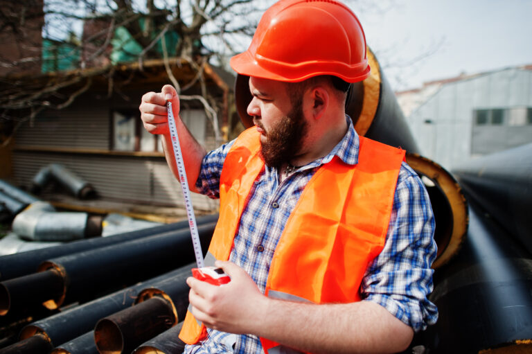 brutal beard worker man suit construction worker safety orange helmet near steel pipes with tape measure hand